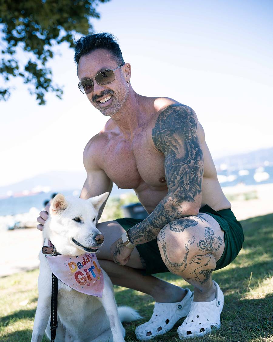 David Turnbull crouching and smiling beside his white dog Juhee, who wears a pink Daddy's Girl bandana, on the grass by a Vancouver beach