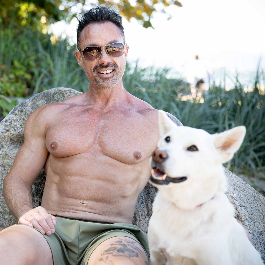 David Turnbull in sunglasses leaning back on a boulder with his white dog Juhee beside him on a Vancouver beach