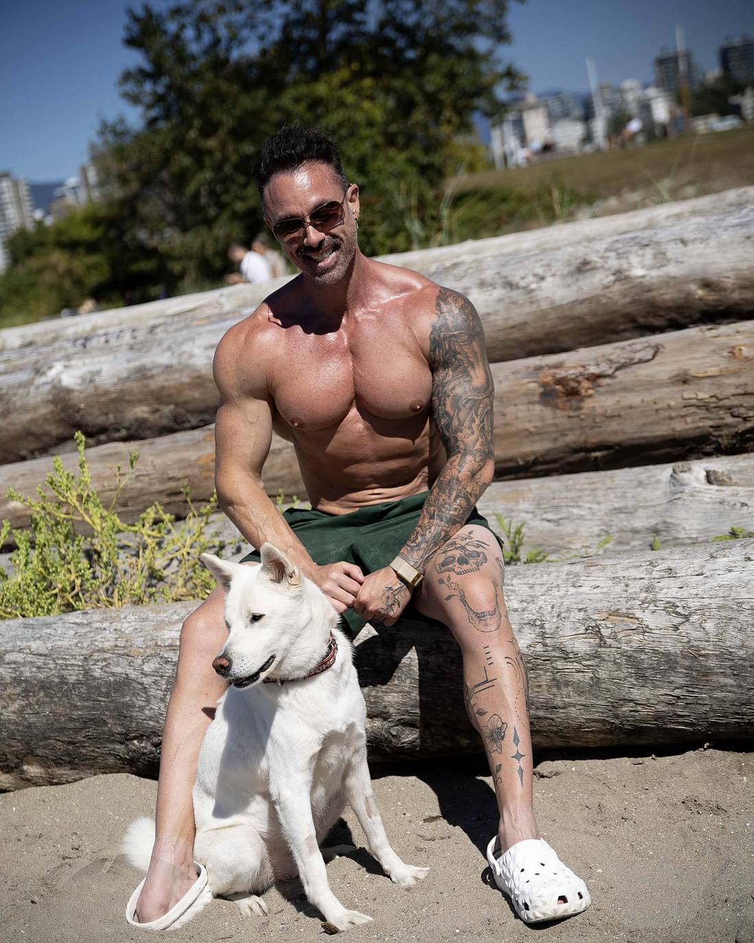 David Turnbull, shirtless and tattooed in sunglasses, sitting on driftwood logs with his white dog on a Vancouver beach